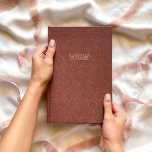 May include: A maroon-colored planner with the text "REAWAKEN PLANNER" is held by two hands. The planner has a leaf pattern and is on a white and pink fabric background.