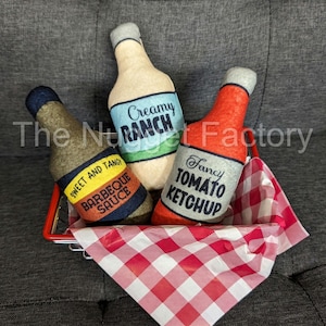 May include: Three felt bottle-shaped pillows in a red wire basket with a red and white checkered cloth. The bottles are labeled "Creamy Ranch", "Fancy Tomato Ketchup", and "Sweet and Tangy Barbeque Sauce".