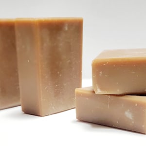 May include: Four bars of brown soap, stacked in a pyramid shape. The soap is a light brown color and has a slightly rough texture.