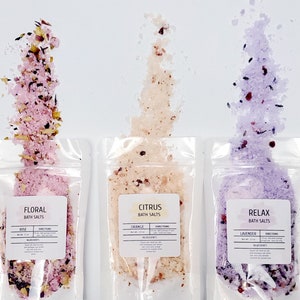 Puede incluir: Tres bolsas de plástico transparentes llenas de sales de baño. Las bolsas están etiquetadas como "Floral Bath Salts", "Citrus Bath Salts" y "Relax Bath Salts". Las sales son de color rosa, naranja y morado, respectivamente. Cada bolsa tiene una pequeña cantidad de flores y hierbas secas.