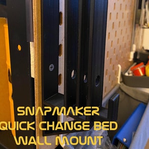 May include: Black 3D printed wall mount for a Snapmaker quick change bed. The mount has a hook on the bottom and is attached to a pegboard.