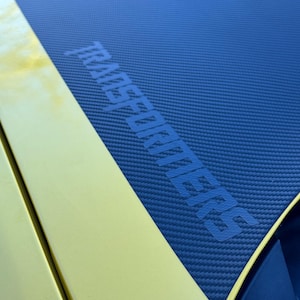 Puede incluir: Primer plano del capó de un coche con una combinación de colores amarillo y azul. La sección azul tiene una textura de fibra de carbono y la palabra "TRANSFORMERS" en azul. La sección amarilla está en el lado izquierdo.