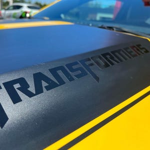 Peut inclure: Gros plan sur le capot jaune d'une voiture avec une section centrale noire. Le mot "TRANSFORMERS" est affiché en grandes lettres noires. Deux bandes jaunes longent les bords du capot.