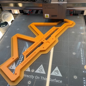 May include: An orange 3D printed quick change bed for a 3D printer. The bed is labeled "The Original Quick Change Bed" and "Laser Version". The text "Front Ant" is also visible on the bed. The bed is sitting on a gray surface with a warning sign that says "Directly On This Surface".