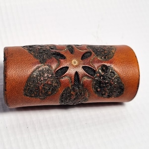May include: A brown leather cuff with a black floral design. The design is intricate and detailed, with a star in the center.
