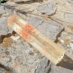 May include: A long, clear rectangular block of resin with embedded orange and brown flecks. The resin is translucent and has a slight shimmer.