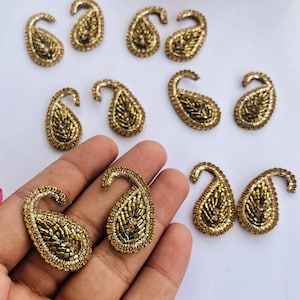 May include: Gold paisley-shaped embellishments with detailed beadwork. The teardrop designs are arranged on a white surface, with some held in a hand. These embellishments are likely for crafting or decorating clothing.