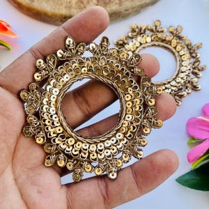 Può includere: Due abbellimenti circolari color oro con intricati dettagli di paillettes e perline. Gli abbellimenti hanno un design floreale sui bordi e sono tenuti in una mano. Lo sfondo include una superficie di legno e fiori colorati.
