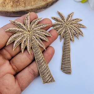 Könnte beinhalten: Zwei gold- und beige-farbene, gestickte Palmen-Applikationen. Die Palmwedel sind hellbeige und die Stämme sind dunkler goldfarben. Die Applikationen befinden sich auf einer weißen Oberfläche.