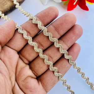 May include: Close-up of gold-colored, wavy trim. The trim has a zig-zag pattern and is held in a hand. The background includes white and red flowers. This trim is suitable for various craft and sewing projects.