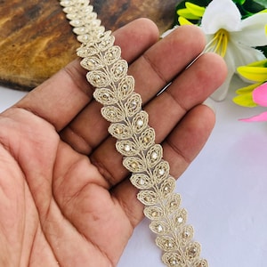 May include: A gold-colored, decorative trim with a leaf-like pattern. The trim is embellished with small, sparkling beads. The trim is held in a hand, with flowers in the background.
