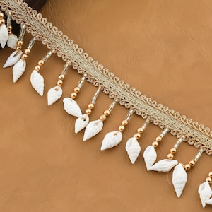 Peut inclure: Garniture dorée et blanche avec une frange perlée et des accents de coquillages blancs.
