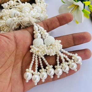 Peut inclure: Pompon de perles ivoire avec un groupe de petites perles et de grosses perles rondes. Le pompon est tenu dans une main, avec un fond en bois et des fleurs blanches.