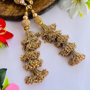 Pode incluir: Close-up de borlas douradas com contas. As borlas são feitas de pequenas contas brilhantes e são presas a uma corrente de ouro. As borlas são dispostas sobre uma superfície de madeira com flores ao fundo. As borlas são provavelmente usadas para joias ou fins decorativos.