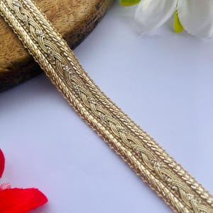 Op de afbeelding: Goudkleurige decoratieve bies met een gevlochten ontwerp. De bies is gemaakt van een glanzend, metallic materiaal en wordt diagonaal tegen een witte achtergrond weergegeven. De bies is ongeveer 2,5 cm breed.