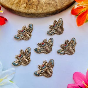 Pode incluir: Seis apliques de abelha bordados. As abelhas têm corpos dourados, asas azuis e detalhes vermelhos. Estão dispostas sobre uma superfície branca, com uma tigela de madeira e flores coloridas no fundo. Ideais para projetos de artesanato.