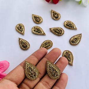 Peut inclure: Appliques dorées en forme de goutte d'eau, ornées de perles. Chaque applique est densément recouverte de petites perles dorées, créant une surface texturée. Certaines sont tenues dans une main, d'autres sont éparpillées sur une surface blanche.