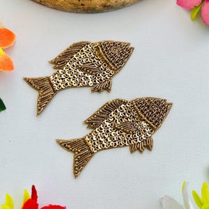 May include: Two gold sequined fish appliques. The fish are intricately detailed with gold beads and sequins, featuring fins, tails, and eye details. The appliques are set against a white background with colourful floral accents.
