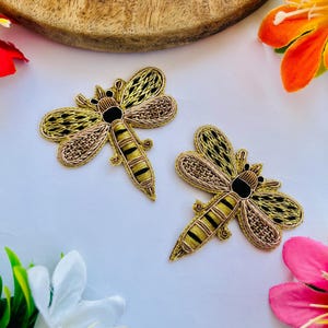 May include: Two embroidered dragonfly patches. The dragonflies feature gold, black, and brown thread detailing. The wings have a delicate, openwork design. The bodies are striped with gold and black. The patches are on a white surface, with flowers and a wooden element in the background.