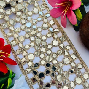 May include: Gold-coloured trim with reflective mirror accents in floral and geometric patterns. The trim is bordered with a delicate gold thread. Pink, red, and white flowers are in the background.