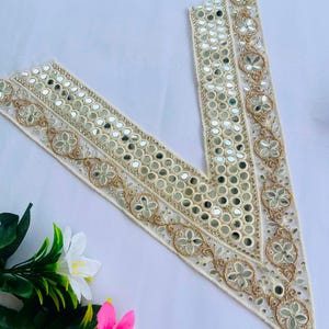 Pode incluir: Acabamento decorativo de cor marfim com pequenos detalhes espelhados redondos e bordados florais dourados. O acabamento tem formato de V, adequado para embelezar roupas ou artesanato. O design apresenta um padrão delicado e ornamentado.