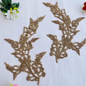 Op de afbeelding: Vier goudkleurige geborduurde bloemenapplicaties met ingewikkelde blad- en bloemontwerpen. De applicaties zijn gerangschikt op een wit oppervlak, met kunstbloemen in de hoeken.