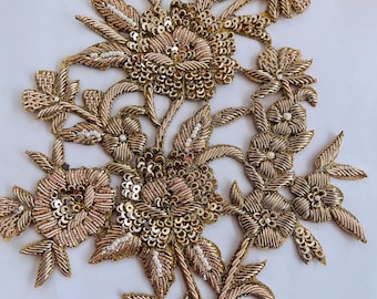 May include: A detailed, gold-colored floral applique with intricate embroidery and sequin embellishments. The design features multiple flower clusters, leaves, and stems, creating a three-dimensional effect. The applique is likely for decorative purposes, such as embellishing clothing or accessories.