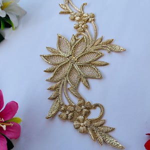 Pode incluir: Aplique floral dourado bordado com detalhes intrincados. O design apresenta uma grande flor aberta, flores menores e folhas delicadas. O aplique é feito com fio metálico e é adequado para embelezar roupas ou artesanato.