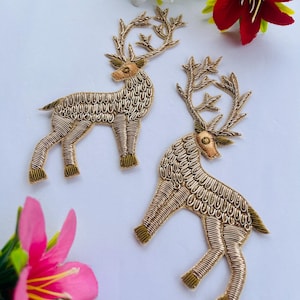 May include: Two gold-colored deer appliqués with intricate embroidery. The deer have detailed antlers and are posed in a standing position. The background is white with colorful flowers.