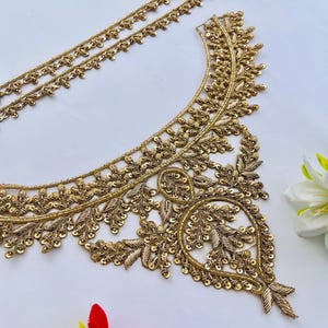 Op de afbeelding: Gouden geborduurde stoffen bies met bloemen- en bladerpatronen. De bies bevat een halslijnontwerp en twee stroken kleinere bies. Het ontwerp is versierd met pailletten en ingewikkelde stiksels. Decoratieve bloemaccenten zijn op de achtergrond.