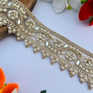 Pode incluir: Borda de renda dourada com detalhes de pérolas e strass. A borda apresenta uma base de rede e uma borda ondulada. O design inclui padrões florais e strass em forma de gota, criando um visual luxuoso. Ideal para artesanato.
