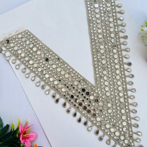 May include: A V-shaped, silver-colored trim with small, round mirrors and teardrop-shaped embellishments. The trim is likely for decorative purposes, possibly for clothing or crafts. The background is white, with a few flowers in the corner.
