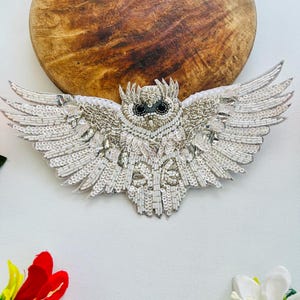 May include: A handcrafted owl applique featuring intricate white and silver beadwork, sequins, and metallic thread embroidery. The owl is depicted with wings fully spread, showcasing detailed plumage textures, black bead eyes, and a pointed beak.