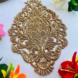 Peut inclure: Applique brodée dorée ornée avec un motif floral symétrique. L'applique présente des détails complexes avec du fil d'or et des paillettes, ressemblant à un arbre ou une plante stylisée. Le fond est blanc, avec des fleurs colorées dans les coins.