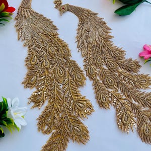 Puede incluir: Dos apliques de pavo real dorados con intrincados detalles de abalorios y lentejuelas. Los apliques presentan patrones de plumas detallados sobre un fondo blanco. Cada pavo real tiene una corona y un ojo rojo, lo que añade elegancia.