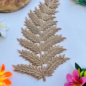 Puede incluir: Una detallada hoja de helecho bordada en color dorado. La hoja está intrincadamente cosida con un hilo metálico, mostrando un patrón detallado. El helecho se coloca sobre una superficie blanca, con coloridas flores artificiales en el fondo.