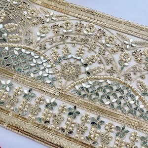 Pode incluir: Uma fita de tecido decorativa com bordado dourado intrincado, pequenas pérolas brancas e peças de espelho refletoras dispostas num padrão floral e arqueado sobre uma base de malha transparente com uma borda dourada nas extremidades.