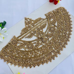 Peut inclure: Applique en dentelle brodée de couleur or, ornée de motifs floraux et géométriques. L'applique est ornée de paillettes et de perles, créant un design décoratif. La dentelle est disposée sur une surface blanche, avec des fleurs colorées en arrière-plan.