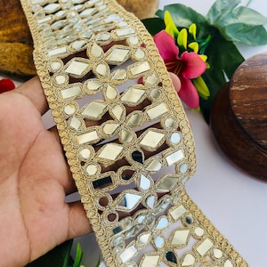 Puede incluir: Ribete decorativo dorado con espejos incrustados. El ribete presenta un patrón repetitivo de formas geométricas y diseños florales. Los bordes están acabados con un delicado borde de cuentas. El ribete se utiliza probablemente para embellecer ropa o proyectos de artesanía.