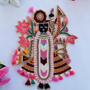 Op de afbeelding: Geborduurde applicatie met een godheid met een zwart gezicht en uitgebreide kleding. Het ontwerp bevat roze, witte en bruine kralen, samen met gouden accenten. Roze kwastjes en bloemmotieven omringen de centrale figuur, waardoor een gedetailleerde en sierlijke uitstraling ontstaat.