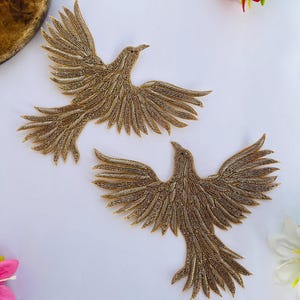 Puede incluir: Dos apliques dorados con forma de pájaro con alas extendidas, elaborados con intrincado trabajo de abalorios. Los pájaros están colocados sobre una superficie blanca, con una flor roja y una flor rosa. Estos apliques son adecuados para adornar ropa o proyectos de manualidades.