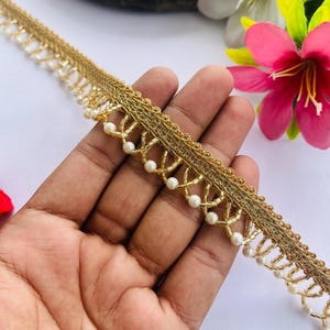 Op de afbeelding: Gouden decoratieve sierrand met een kralenrand. De rand heeft een geweven ontwerp met kleine, witte, parelachtige kralen. De rand wordt in een hand gehouden, met bloemen op de achtergrond.