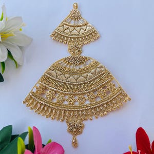May include: A gold-coloured, ornate, tiered hair ornament with a teardrop-shaped pendant. The ornament is made of metal and has a delicate, intricate design.