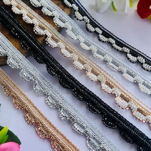 May include: Close-up of several decorative trims in various colors. The trims feature a scalloped edge with beaded detailing. Colors include black, silver, gold, and white. The trims are arranged diagonally, showcasing their design and texture. Ideal for embellishing garments or crafts.