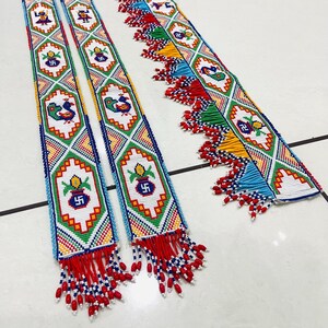 Puede incluir: Dos guirnaldas de cuentas con patrones geométricos coloridos y borlas. Las guirnaldas presentan un diseño repetitivo de pavos reales, macetas y símbolos de esvástica.