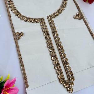 May include: A white fabric with intricate gold floral embroidery. The embroidery outlines a neckline and a central panel, with additional floral accents. The design is likely for a garment, showcasing detailed craftsmanship and a decorative aesthetic.