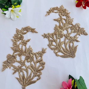 Puede incluir: Dos apliques florales bordados en color oro. Los apliques presentan diseños detallados de flores y hojas. El bordado está realizado con un hilo metálico, lo que da a los apliques un aspecto brillante. Los apliques están colocados sobre un fondo blanco.