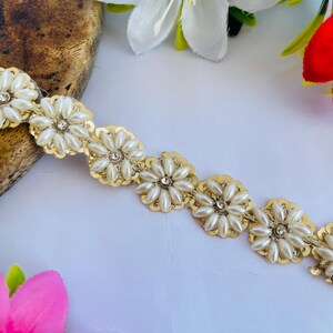 May include: A decorative trim featuring a repeating floral pattern. Each flower has a gold-colored scalloped edge with a cluster of faux pearls and a central rhinestone. The trim is on a white background with flowers.