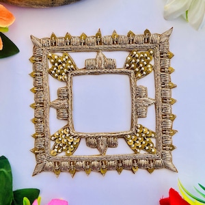 May include: A square-shaped gold embroidered patch featuring intricate metallic thread work, raised corded detailing, and small gold sequins. The design includes a central open square frame surrounded by decorative leaf-like motifs and beaded accents.