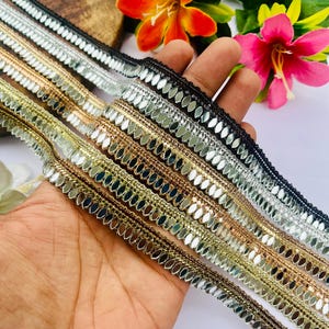 May include: A collection of five decorative fabric trims featuring rows of small, oval-shaped reflective mirror pieces. The trims are arranged in a fan shape, showcasing black, silver, copper, gold, and bronze metallic thread borders.